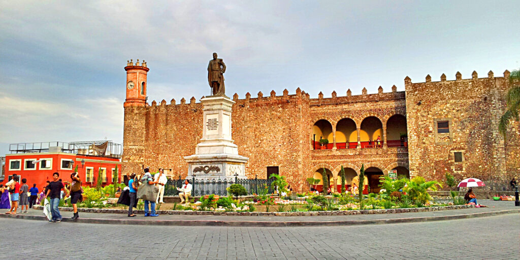 Palacio de Cortés en Cuernavaca, México, con una estatua frente al edificio rodeada de jardines y personas caminando en la plaza