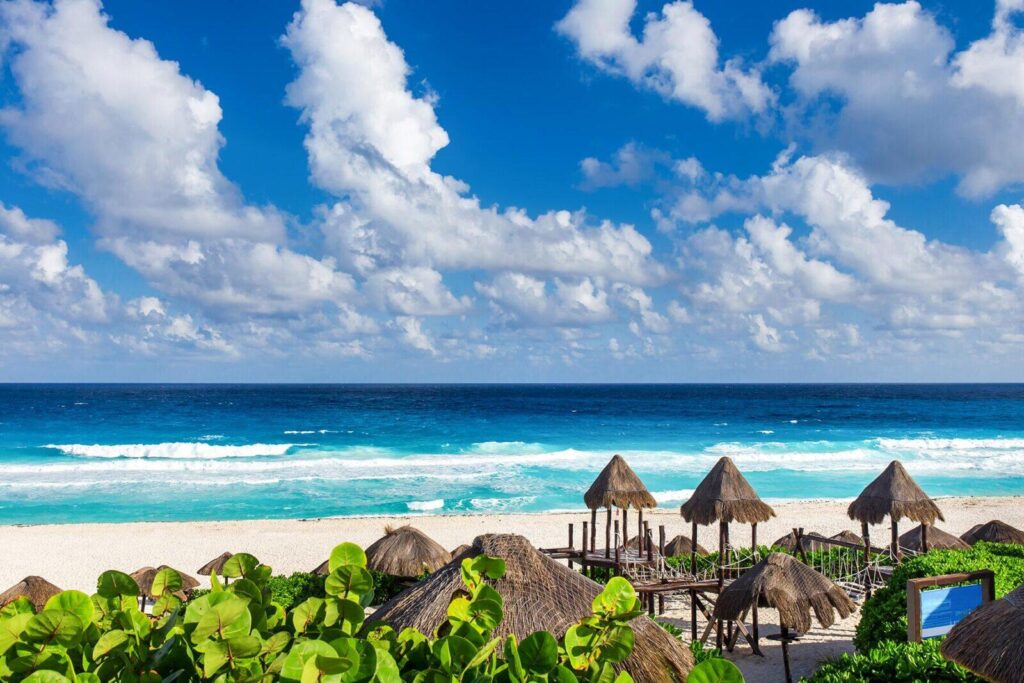 Playa tropical con arena blanca, agua turquesa y palapas con techos de palma frente al mar bajo un cielo azul con nubes dispersas