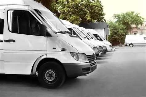 Fila de camionetas de carga blancas estacionadas en un lote, alineadas de manera ordenada, con árboles y edificios al fondo.